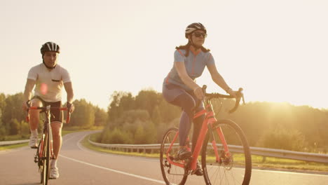 Steadicam-shot-of-two-healthy-mem-and-woman-peddling-fast-with-cycling-road-bicycle-at-sunset